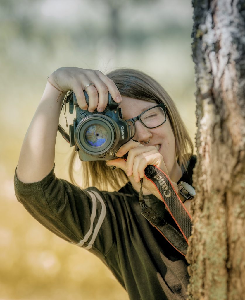 Frau fotografiert hinter Baum hervor, Fotografin
